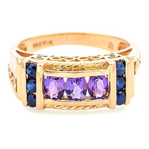 May include: A gold ring featuring three oval-cut purple gemstones in the center, flanked by three smaller blue gemstones on each side. The ring has a decorative, textured design.