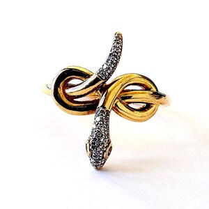 May include: A gold-toned ring in the shape of a snake, with a coiled body and a head adorned with small, sparkling stones. The snake's head features tiny red accents, and the ring is set against a white background.
