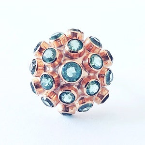 May include: A rose gold ring with a cluster of teal-colored gemstones. The ring's design features multiple round gemstones set in a spherical arrangement. The gemstones are a vibrant teal color, contrasting with the warm rose gold setting. The ring is set against a white background.