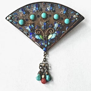 May include: An ornate fan-shaped brooch with turquoise and red accents. The metalwork features intricate blue and gold enamel designs, including a butterfly motif. The brooch has a dangling charm with turquoise and red teardrop-shaped beads.