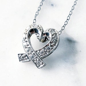May include: A silver-toned heart pendant necklace with a delicate chain. The heart is encrusted with small, sparkling stones and features a ribbon-like design. The pendant is set against a white background.