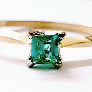 May include: A gold ring featuring a square-cut, vibrant green emerald gemstone. The emerald is set in a silver-colored prong setting, and the band is a simple, polished gold. The ring is displayed against a white background.