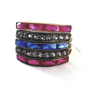 May include: Stack of gold-toned rings with alternating bands of pink and blue gemstones, and bands of small, clear stones. The gemstones are rectangular and faceted. The rings are stacked together on a white background.