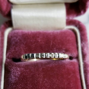 May include: A gold ring with small square-cut stones set in a row. The ring is displayed in a pink velvet jewelry box with a cream-colored interior. The box has text that reads "GRD BOULEVARD AND GRATIOT AVE DETROIT, MICH."