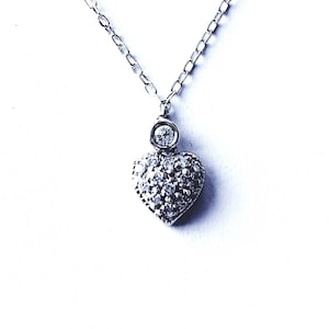 May include: A silver necklace with a heart-shaped pendant. The pendant is encrusted with small, sparkling stones, and a single, larger stone sits above the heart. The chain is delicate and hangs against a white background.