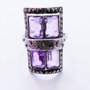 May include: A silver ring featuring two rectangular, light purple gemstones. The ring has a decorative band with a pattern of small, dark stones around the gemstones. The ring is set against a white background.