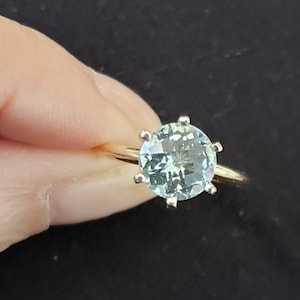 May include: A gold ring featuring a large, round, light blue gemstone. The gemstone is held in place by six prongs. The ring is set against a dark background.