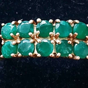 May include: A gold-toned ring featuring two rows of round, vibrant green gemstones. The gemstones are evenly spaced and set in a gold band, creating a symmetrical and elegant design. The ring is displayed against a dark background.