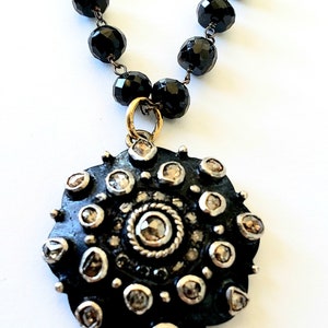 May include: A black beaded necklace with a large, round pendant. The pendant is dark with a silver border and small, round, clear stones. The necklace has a gold-colored clasp.