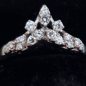 May include: A silver ring with a crown-like design, set with multiple clear, sparkling gemstones. The ring features a central, pointed cluster of stones, flanked by smaller, rounded clusters, creating a delicate and elegant appearance. The ring is set against a dark background.
