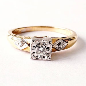 May include: A gold ring with a square, ornate, white gold setting holding a single, round diamond. The band has diamond-shaped accents with small diamonds. The ring is set against a white background.