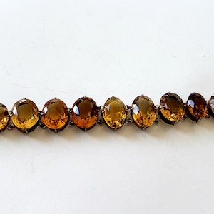May include: A bracelet featuring oval-shaped amber-colored gemstones set in a dark metal frame. The gemstones graduate in size, creating a visually appealing design. The bracelet is displayed on a white surface.