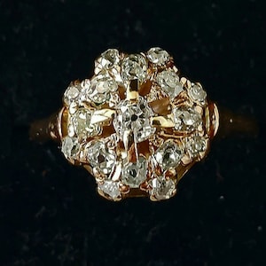May include: A gold ring featuring a cluster of small, clear gemstones. The gemstones are arranged in a floral or starburst pattern, set against a gold band. The ring is displayed against a dark background.