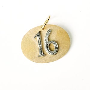 May include: A gold-toned pendant with the number "16" in silver, encrusted with small, sparkling stones. The pendant has a textured, oval shape and a gold loop for hanging. The background is white.