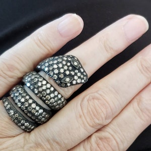 May include: A silver snake ring with a head and body that wraps around the finger. The ring is covered in small, sparkling stones, with two oval-shaped stones for eyes. The snake's head is slightly raised, giving it a three-dimensional appearance.