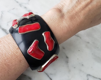 May include: A wide, black cuff bracelet with red and silver accents. The bracelet features several irregularly shaped red inlays set in silver. The bracelet is worn on a wrist, showcasing its bold design and contrasting colors.
