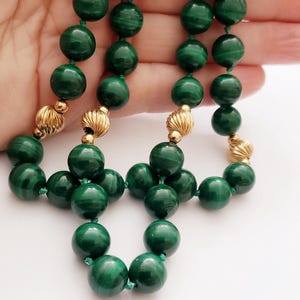 May include: A close-up of a necklace featuring large, round, dark green beads. The beads are strung together with a green cord and separated by gold-colored, textured accents. The necklace is displayed against a light background.