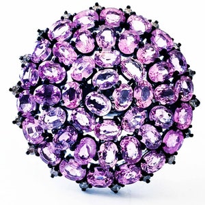 May include: A round, ornate ring featuring numerous oval-cut, light purple gemstones. The gemstones are set closely together, creating a clustered, floral-like design. The ring's setting is dark, possibly black, with small accent stones around the edges.