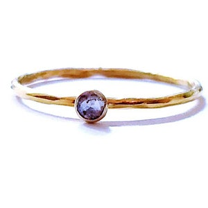 May include: A delicate gold ring featuring a small, round, light purple gemstone. The ring has a thin band with a hammered texture, and the gemstone is set in a simple bezel. The ring is displayed on a white background.