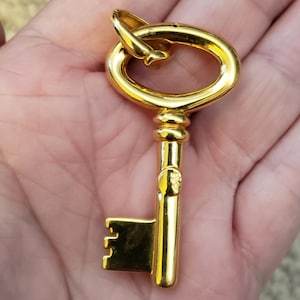 May include: A gold-colored key pendant with a classic design. The key has a rounded top, a detailed stem, and a rectangular key bit. The pendant is held in a hand, showcasing its size and shine. This jewelry item is suitable for necklaces or keychains.