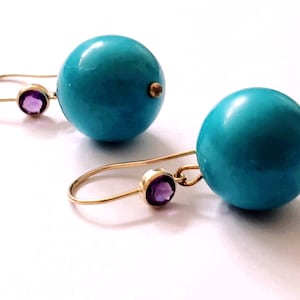 May include: A pair of turquoise blue ball earrings with gold-colored hooks. Each earring features a small, round, purple gemstone accent. The earrings are set against a white background.