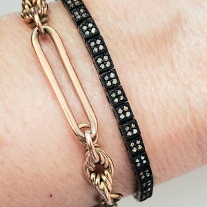 May include: Two bracelets are shown. One is a gold-colored link bracelet with a decorative clasp. The other is a black bracelet with small, square, silver-colored accents. The bracelets are worn together.