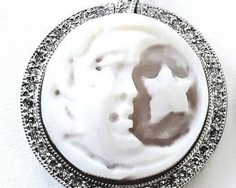 May include: A round pendant with a white cameo featuring a profile and star design. The pendant is set in a silver-toned frame with a border of small, clear gemstones. The bail is also adorned with gemstones.