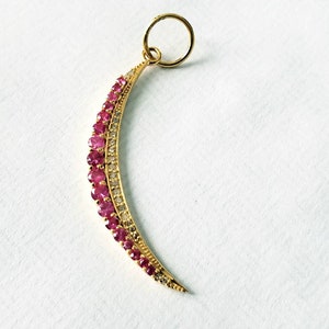 May include: A crescent-shaped pendant with a gold-colored metal frame. The pendant is adorned with rows of small, bright red gemstones and smaller, clear gemstones. A gold-colored loop is at the top for hanging.