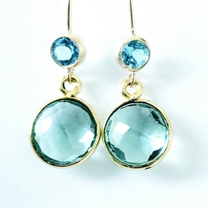 May include: A pair of gold-toned drop earrings. Each earring features a small, round, light blue gemstone at the top and a larger, round, light blue gemstone at the bottom. The gemstones are set in gold-colored metal.