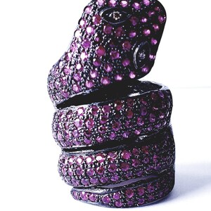 May include: A unique, dark metal snake ring with a coiled design. The ring is encrusted with numerous small, round, magenta-colored gemstones, creating a sparkling effect. The snake's head is raised, adding a distinctive touch.