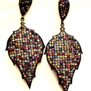 May include: A pair of ornate earrings with a leaf-like design. The earrings feature a dark metal frame and are encrusted with a mosaic of colorful gemstones in shades of red, yellow, blue, and purple. The earrings have a teardrop-shaped top.