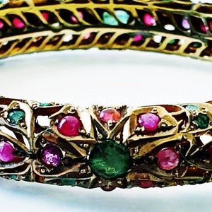 May include: Ornate gold-toned bangle bracelet. The bracelet is encrusted with vibrant red and green gemstones, with a central green stone. The design features intricate patterns and a detailed, vintage aesthetic. A piece of jewelry.