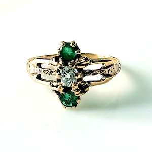 May include: A gold ring featuring a central diamond flanked by two emeralds. The ring has an ornate design with decorative cutouts and a polished finish. The gemstones are set in a prong setting.