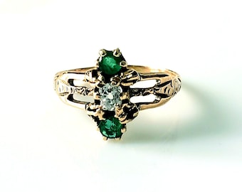 May include: A gold ring featuring a central diamond flanked by two emeralds. The ring has an ornate design with decorative cutouts and a polished finish. The gemstones are set in a prong setting.