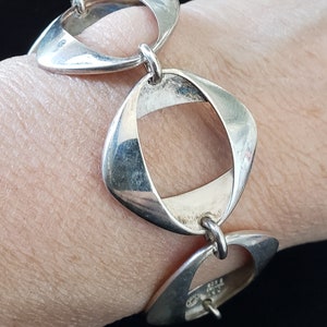 May include: A silver bracelet featuring three open, geometric shapes linked together. The bracelet is worn on a wrist, showcasing its design and craftsmanship. The shapes are slightly curved and have a polished finish.