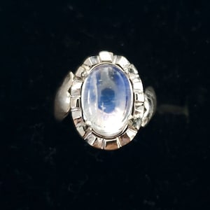 May include: A silver ring with an oval, iridescent moonstone. The stone is set in a decorative bezel with a detailed design. The ring has a smooth band and is displayed against a black background.