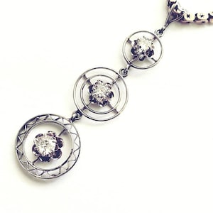 May include: A silver necklace with three circular pendants, each featuring a central clear gemstone. The pendants are connected by delicate links, and the necklace has a chain with small, star-shaped links. The design is intricate and elegant.