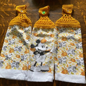 Op de afbeelding: Drie gele gehaakte theedoeken met een Mickey Mouse- en pompoenprint. De handdoeken hebben een knoop aan de bovenkant.