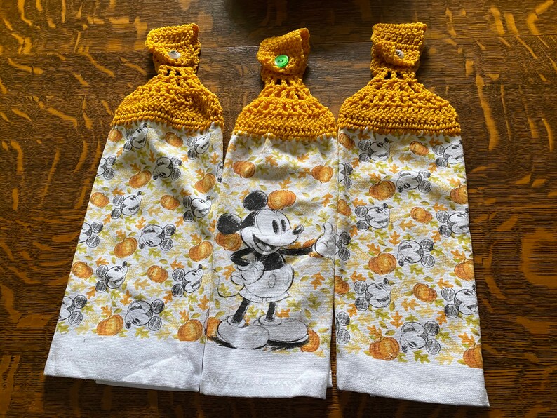 Op de afbeelding: Drie gele gehaakte theedoeken met een Mickey Mouse- en pompoenprint. De handdoeken hebben een knoop aan de bovenkant.