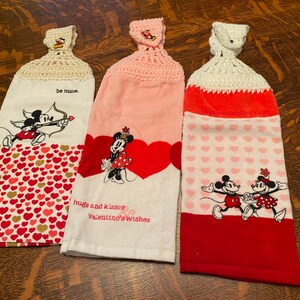 Op de afbeelding: Drie theedoeken met Valentijnsdagthema met Mickey Mouse en Minnie Mouse-ontwerpen. De theedoeken zijn wit met rode en roze harten en hebben gebreide toppen. De tekst op de theedoeken luidt "be mine", "hugs and kisses Valentine's wishes" en "Happy Valentine's Day".