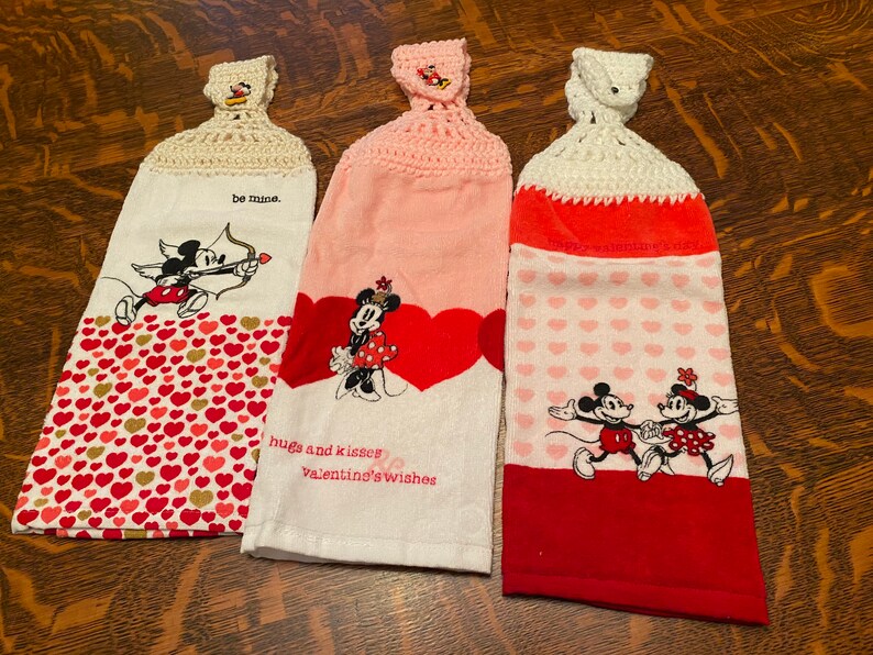 Op de afbeelding: Drie theedoeken met Valentijnsdagthema met Mickey Mouse en Minnie Mouse-ontwerpen. De theedoeken zijn wit met rode en roze harten en hebben gebreide toppen. De tekst op de theedoeken luidt "be mine", "hugs and kisses Valentine's wishes" en "Happy Valentine's Day".