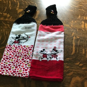 May include: Two Valentine's Day themed kitchen towels with Mickey Mouse and Minnie Mouse designs. The towels have black crocheted hanging loops with Mickey Mouse heads. One towel has a red and white heart pattern and says "be mine." The other towel has a red, white, and pink heart pattern and shows Mickey and Minnie holding hands.