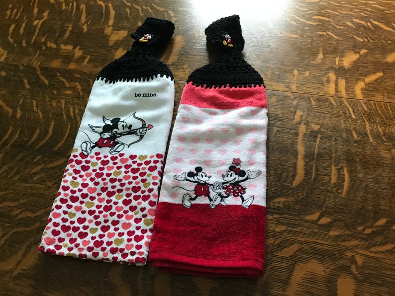 May include: Two Valentine's Day themed kitchen towels with Mickey Mouse and Minnie Mouse designs. The towels have black crocheted hanging loops with Mickey Mouse heads. One towel has a red and white heart pattern and says "be mine." The other towel has a red, white, and pink heart pattern and shows Mickey and Minnie holding hands.