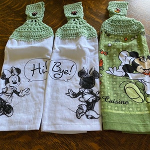 Op de afbeelding: Drie keukendoeken met groene gehaakte toppen. De handdoeken hebben verschillende Disney-figuren: Mickey Mouse, Minnie Mouse en Minnie Mouse. De handdoeken hebben de tekst "Hi! Bye!" en "Cuisine" erop gedrukt.