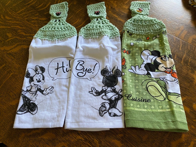 Op de afbeelding: Drie keukendoeken met groene gehaakte toppen. De handdoeken hebben verschillende Disney-figuren: Mickey Mouse, Minnie Mouse en Minnie Mouse. De handdoeken hebben de tekst "Hi! Bye!" en "Cuisine" erop gedrukt.