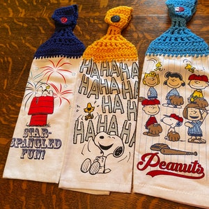 Op de afbeelding: Drie gehaakte theedoeken met Peanuts-figuren. De eerste theedoek heeft Snoopy en Woodstock met vuurwerk en de tekst "Star-Spangled Fun!" De tweede theedoek heeft Snoopy die lacht en de tekst "HAHAHA HA HA HA HA HA HA". De derde theedoek heeft een honkbal sc&egrave;ne met Snoopy en de tekst "Peanuts".