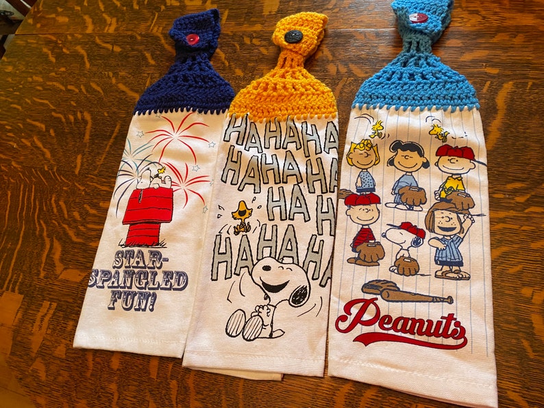 Op de afbeelding: Drie gehaakte theedoeken met Peanuts-figuren. De eerste theedoek heeft Snoopy en Woodstock met vuurwerk en de tekst "Star-Spangled Fun!" De tweede theedoek heeft Snoopy die lacht en de tekst "HAHAHA HA HA HA HA HA HA". De derde theedoek heeft een honkbal sc&egrave;ne met Snoopy en de tekst "Peanuts".