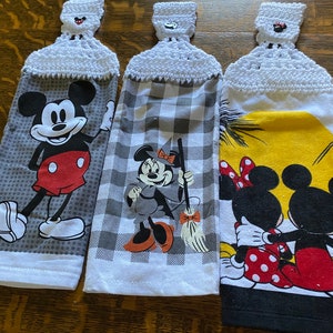 May include: Three hanging kitchen towels with crochet tops. Each towel features a different Mickey Mouse design: Mickey waving, Minnie Mouse as a witch, and Mickey and Minnie Mouse embracing. The towels have white crochet tops.
