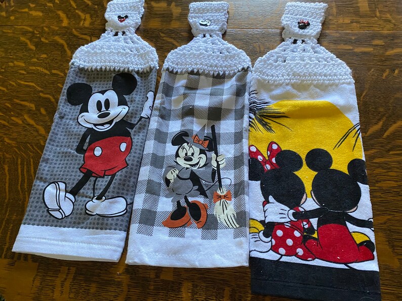 May include: Three hanging kitchen towels with crochet tops. Each towel features a different Mickey Mouse design: Mickey waving, Minnie Mouse as a witch, and Mickey and Minnie Mouse embracing. The towels have white crochet tops.