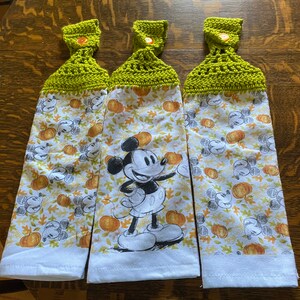 Op de afbeelding: Drie witte keukendoeken met een groene gehaakte bovenkant en een knoop. De handdoeken hebben een Mickey Mouse-figuur en een herfstthema met pompoenen en bladeren.